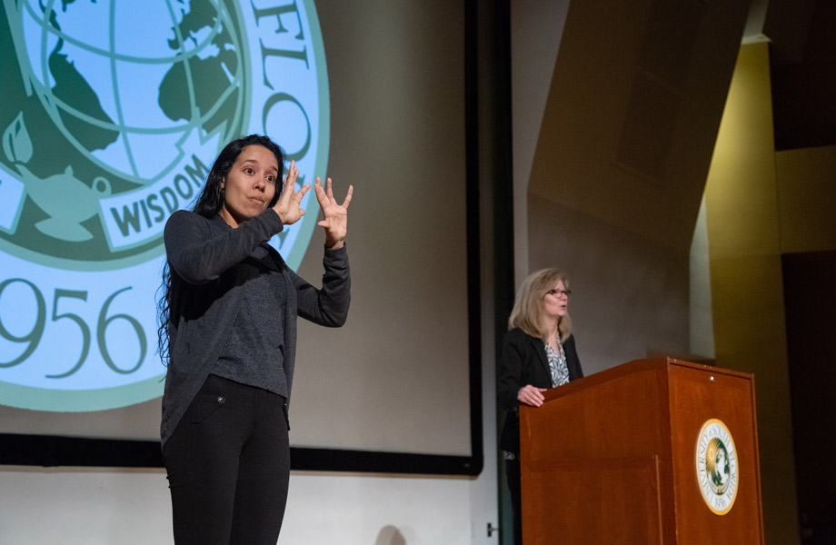 Woman offering ASL interpretation during speech, with ֱ seal behind her